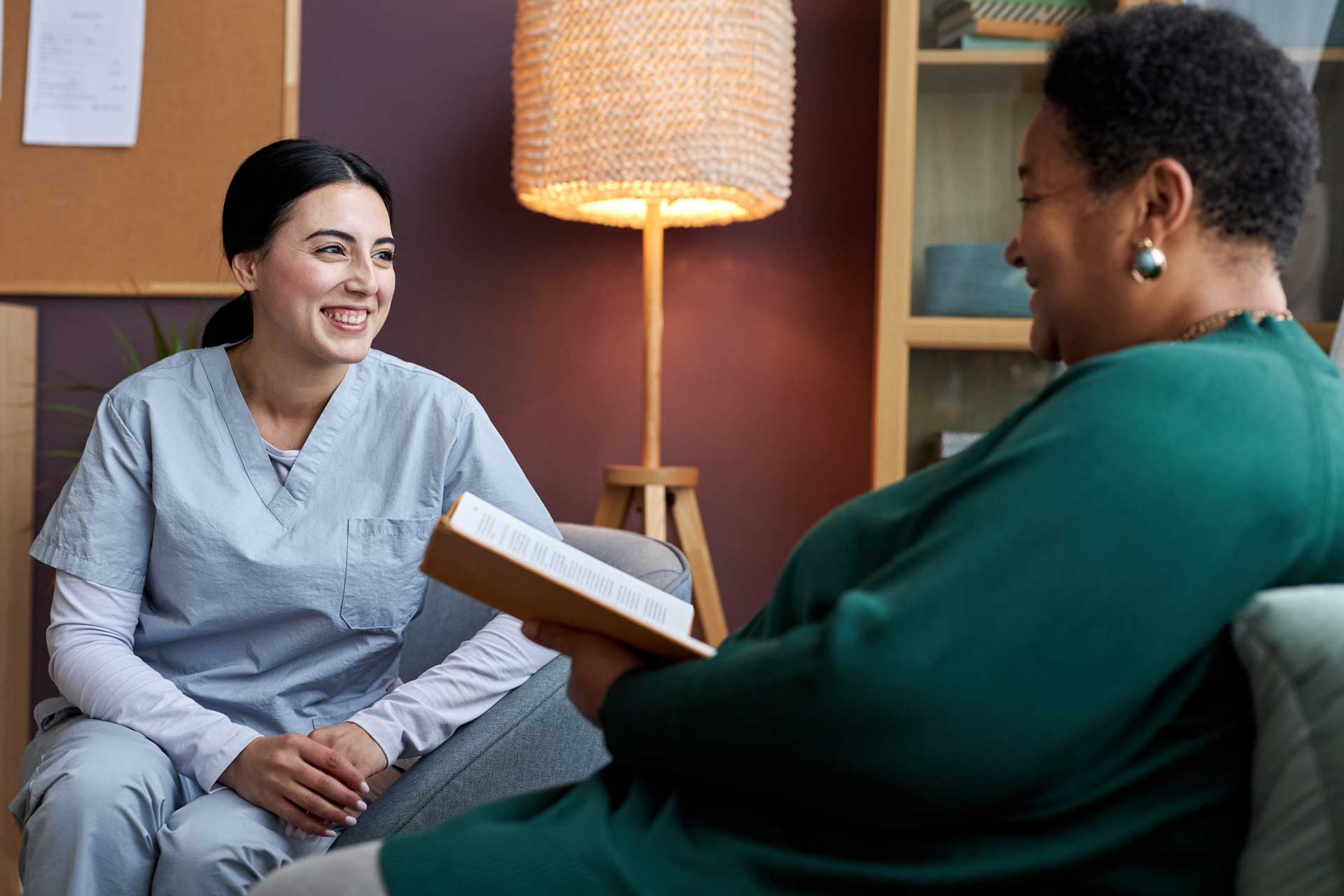 two women smiling at each other, one in scrubs and the other reading a book